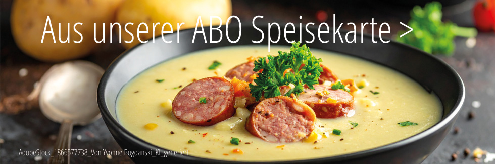 Eine Suppenschüssel mit Kartoffelsuppe und Würstchen garniert mit Petersilie, links ein Löffel und im Hintergrund zwei Kartoffeln