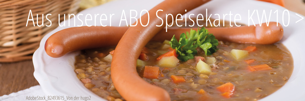 Auf einem Holztisch steht ein weißer Teller mit Linseneintopf. In diesem sehen der Kartoffeln, gelbe Rüben und Petersilie. Über kreuzliegend, zwei Wiener Würstchen.